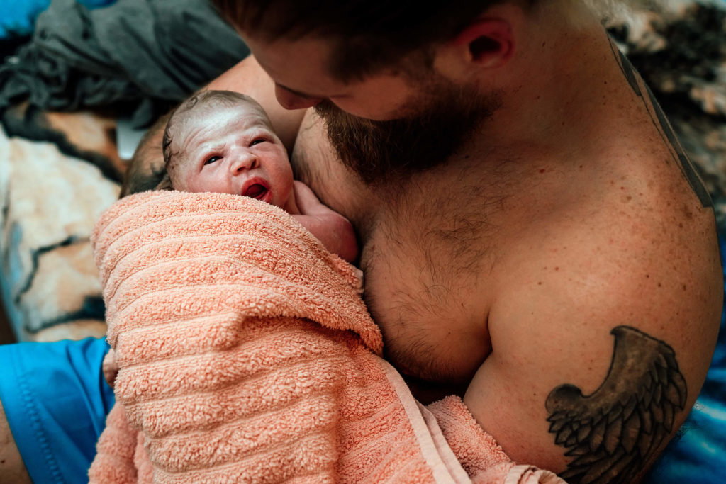 dad holding new baby while midwives assist mom out of the birth tub at home