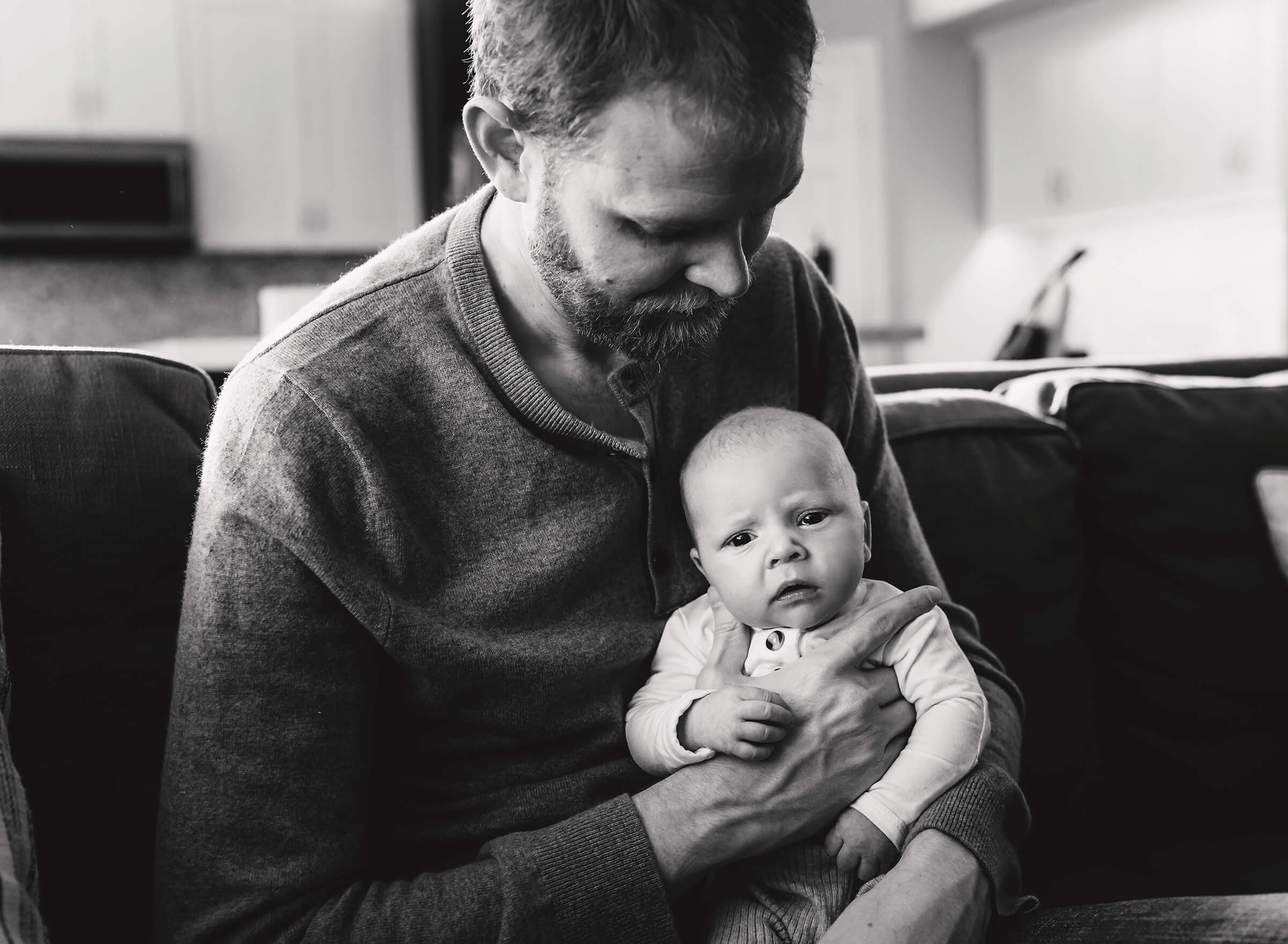 black and white image of dad holding newborn son on his lap at home during photography session 