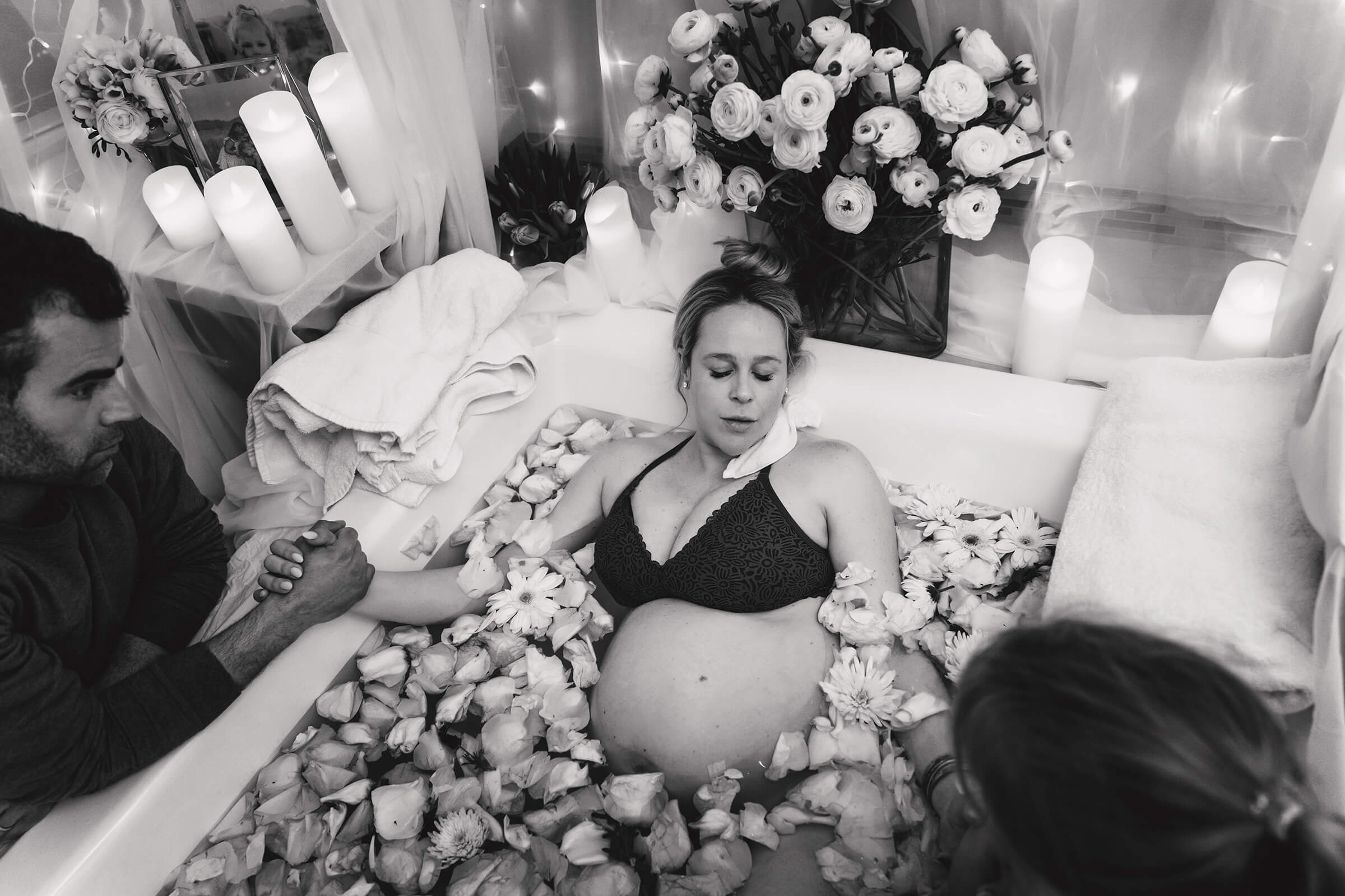 black and white image of a woman laboring in a tub full of petals surrounded by white flowers and beautiful light. 