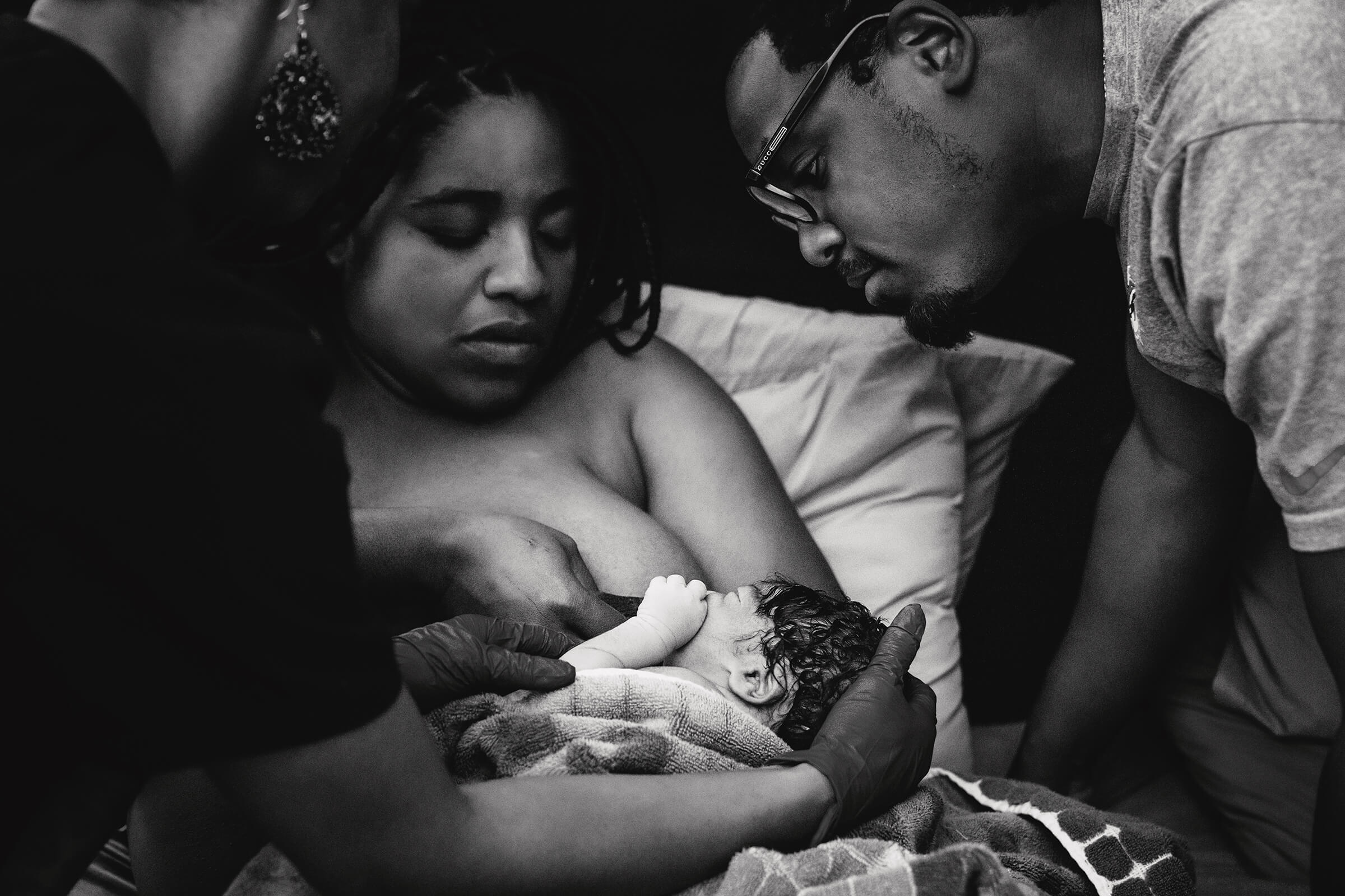 black and white image of a mother breastfeeding her new baby as her husband looks on