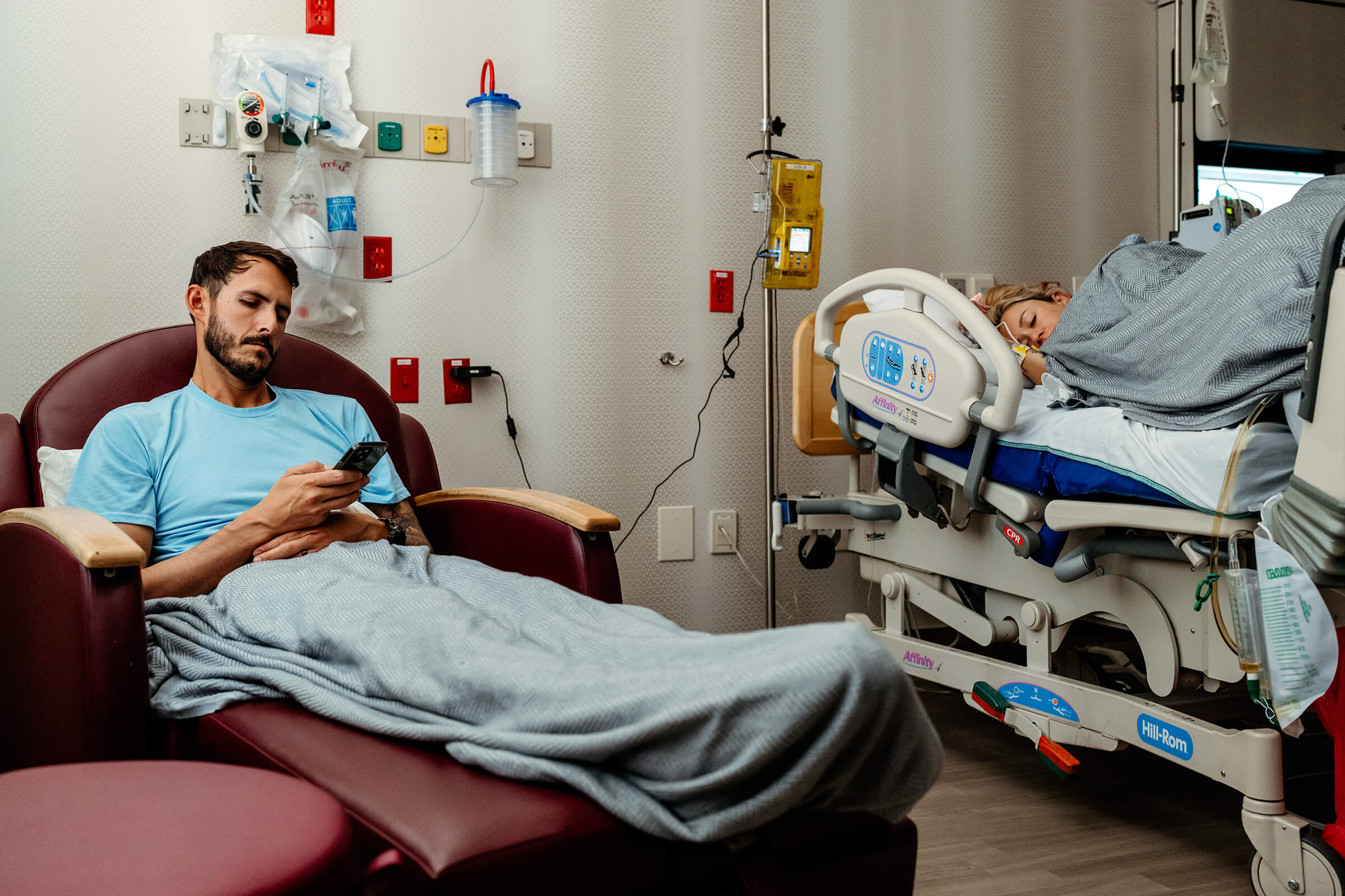 mother in side-lying position in hospital bed while husband updates the family on his phone